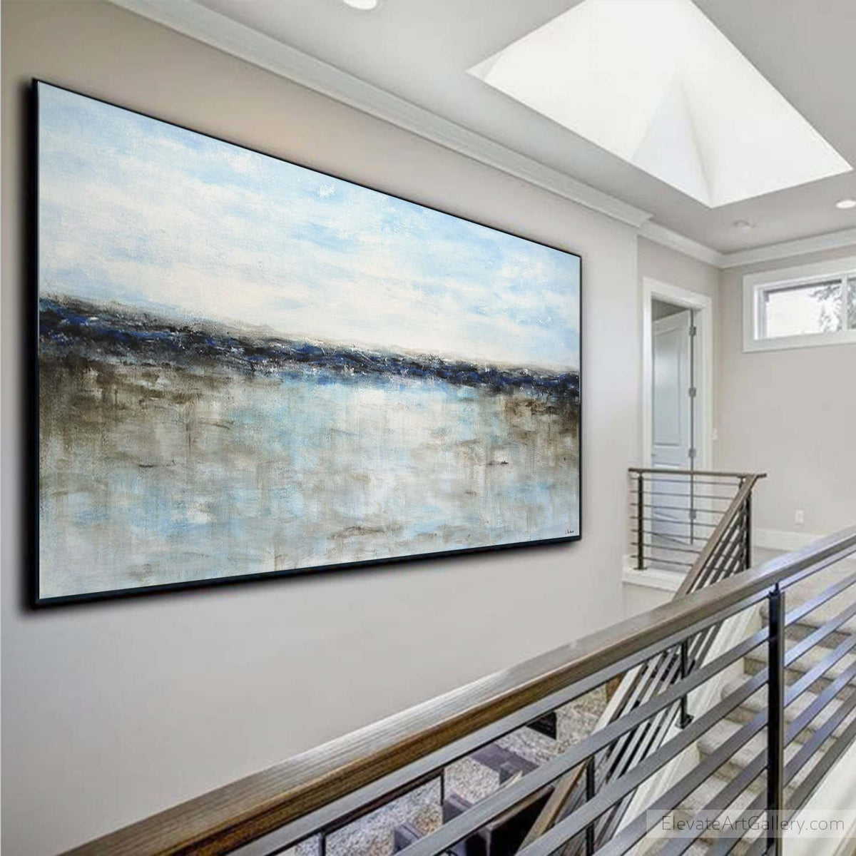 A large framed abstract landscape painting with blue, gray, and white tones hangs on a beige wall in a modern staircase hallway with a wooden and metal railing.